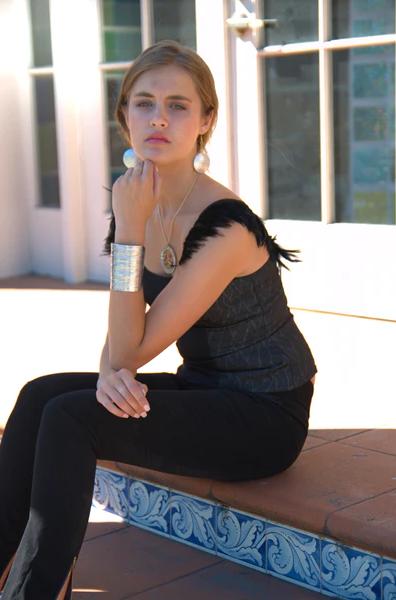 A woman in a black top sitting on a brick wall, exuding a calm and confident demeanor wearing aluminum statement cuff