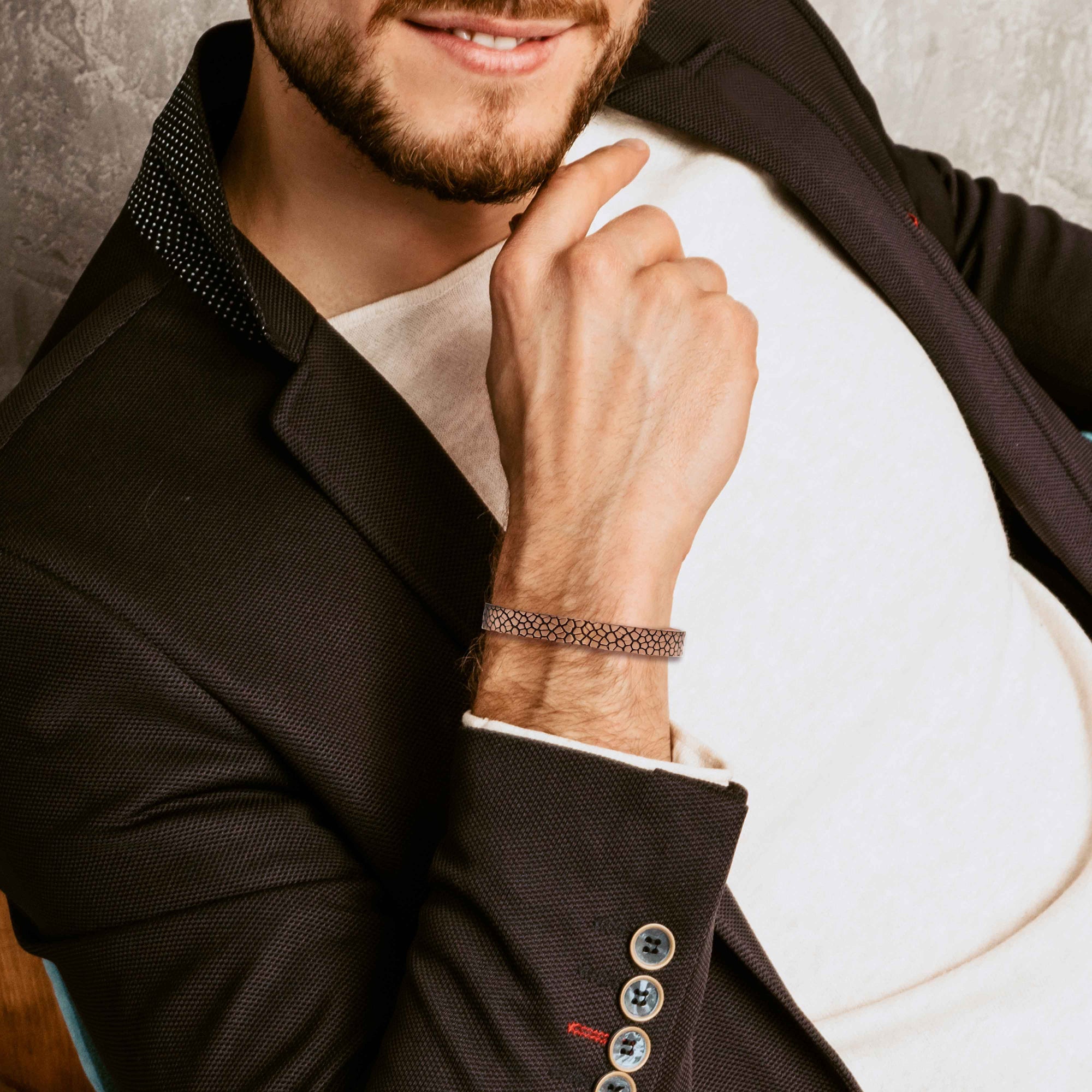 Lifestyle image of a man wearing a slim snakeskin patterned copper cuff bracelet on his wrist, styled with a white shirt and dark textured blazer. The solid copper cuff features a fine, scale-like embossed pattern across the surface, giving it a refined yet rugged texture that contrasts with the clean tailoring of his outfit. The warm copper tone sits close to the wrist, showing how the narrow patterned bracelet adds a subtle, modern accent to a dressed-up, everyday look.