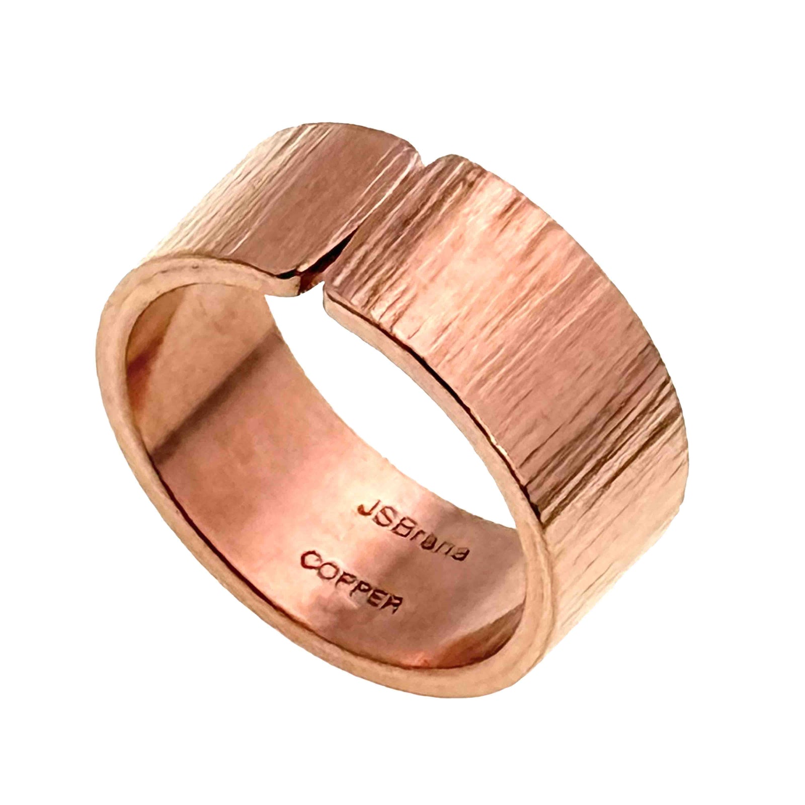 Handmade solid copper cuff ring shown at an angle on a white background. The ring has a wide band with an open back and a hand chased texture across the outside, creating soft linear markings that catch the light. The interior is smooth and polished, stamped with “JSBrana” and “COPPER.” Warm rose copper tones and the bold 10mm width give it a modern, artisan look.