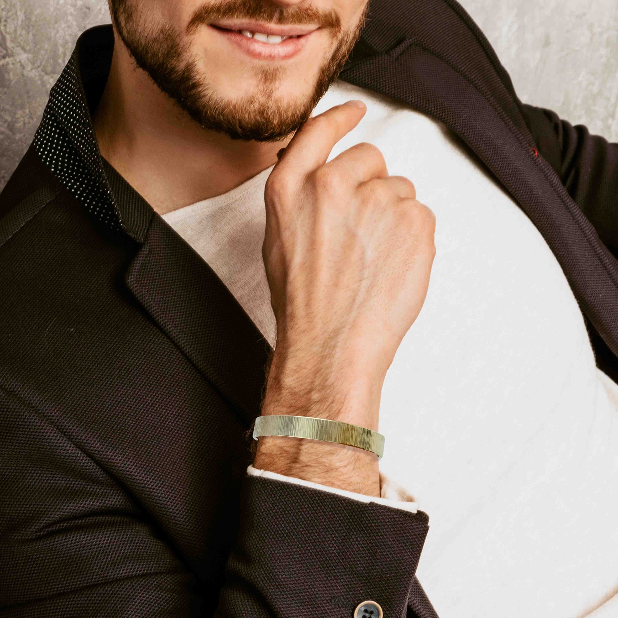 Man wearing a 10mm Wide Chased Nu Gold Tone Brass Cuff Bracelet on his wrist, dressed in a dark suit and white shirt.