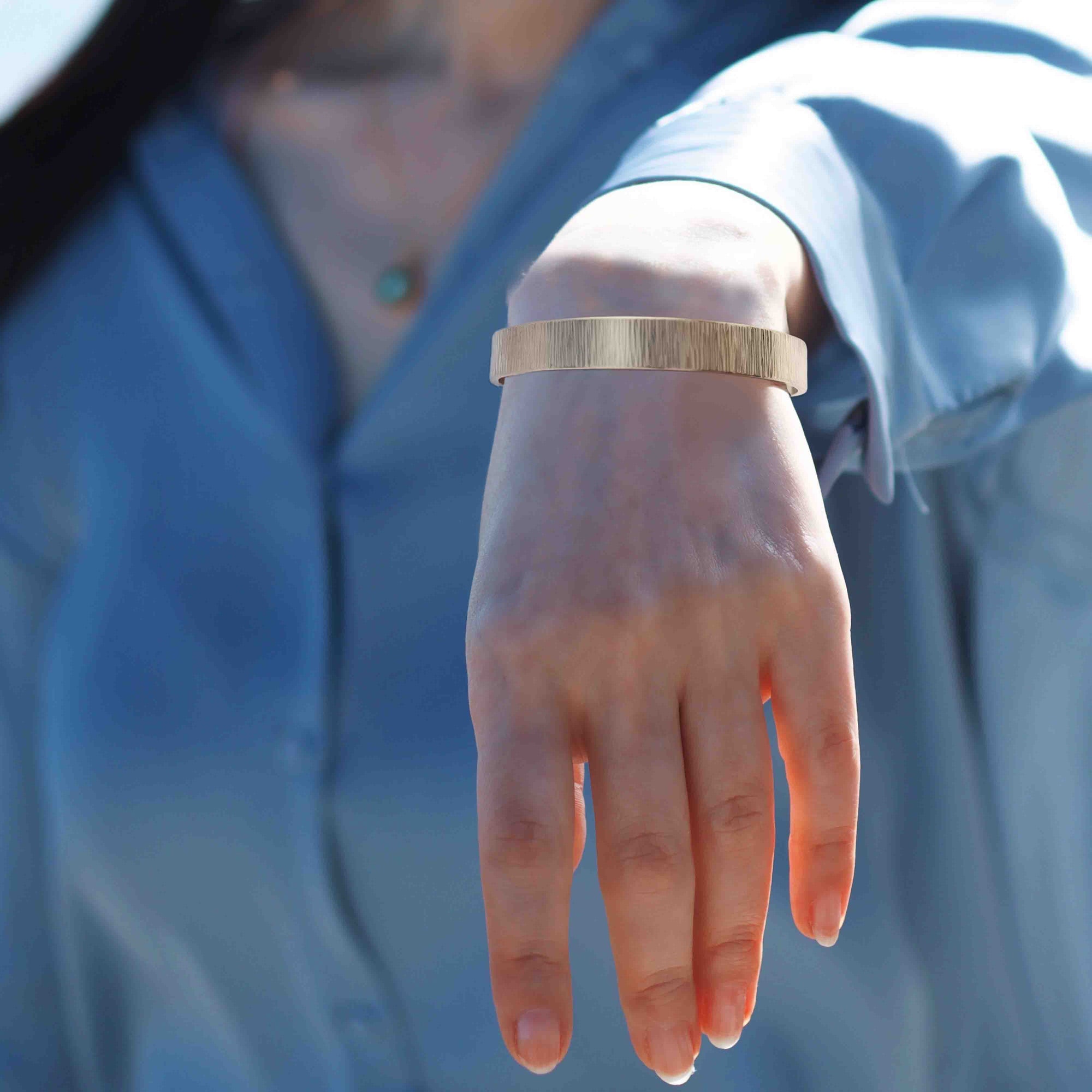 A Woman's wrist wearing a 10mm Wide Chased Nu Gold Tone Brass Cuff Bracelet with a blurred blue background