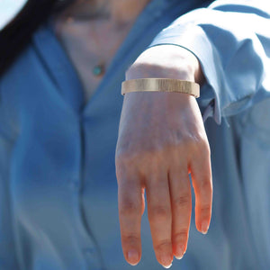 4 of 9: A Woman's wrist wearing a 10mm Wide Chased Nu Gold Tone Brass Cuff Bracelet with a blurred blue background