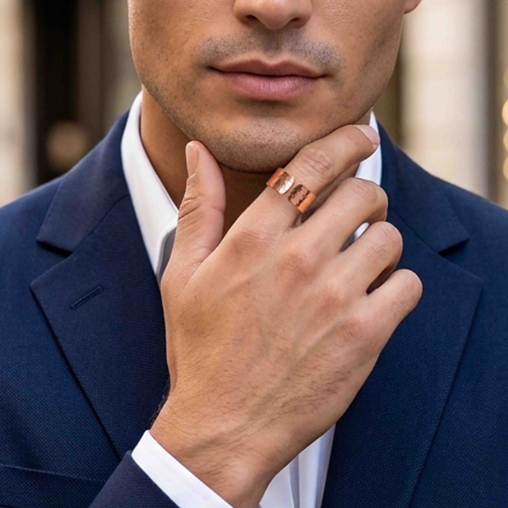 Close-up of a man in a navy suit and white dress shirt wearing a 10mm wide solid copper cuff ring on his finger. The open adjustable ring has a polished hammered finish that catches the light, adding texture and warmth to the bold copper tone. His hand rests at his chin, giving the ring a refined, masculine look against the softly blurred background and tailored formal styling.