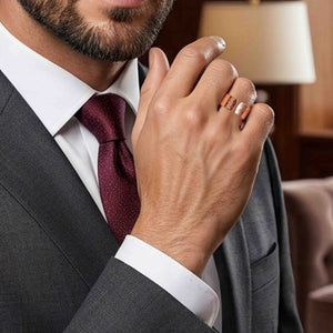 4 of 9: Close up of a man in a gray suit and burgundy tie wearing a wide hammered copper cuff ring on his finger. His hand is raised near his face, highlighting the ring’s polished surface and open cuff design, with warm copper tones contrasting against his formal attire and softly lit indoor background.