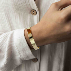4 of 9: Close-up of a woman's wrist wearing a 10mm Hammered Nu Gold Brass Cuff Bracelet on a light-colored shirt.