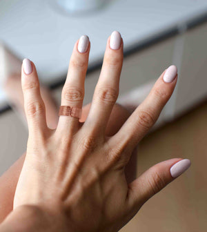 5 of 8: A close-up of a hand with light pink manicured nails wearing a wide, open-front copper cuff ring with a linen-textured finish. The ring has a warm, reddish hue and stands out against the skin tone and soft nail color. The background is softly blurred, suggesting an indoor setting.