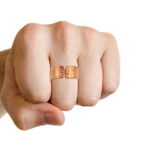 7 of 8: A close-up of a hand wearing a wide copper cuff ring with a linen-textured finish and open-front design. The ring sits on the middle finger, showcasing its warm reddish tone against light skin. The background is plain white, highlighting the ring’s handcrafted detail.