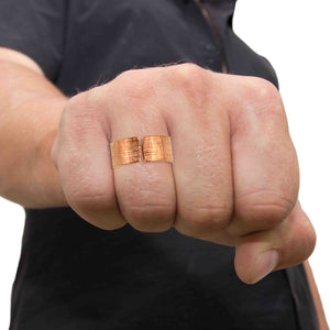 2 of 8: A close-up of a person wearing a wide, open-front linen-textured copper cuff ring on their finger. The ring has a warm, reddish tone and a bold, handcrafted look. The person is clenching their fist, showcasing the ring against a dark shirt and light background.