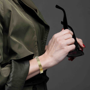 7 of 9: A Woman holding a pair of black sunglasses with a plain background wearing a 10mm Wide Linen Nu Gold Tone Brass Cuff Bracelet on her wrist