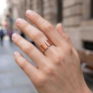 2 of 9: Close-up of a hand wearing a matte copper cuff ring with a wide open band design, featuring a smooth brushed finish and warm reddish tone, worn on the middle finger against a softly blurred city street background.