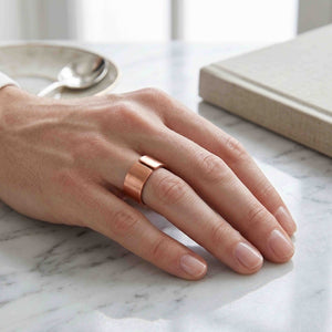 6 of 9: Close-up of a hand resting on a light marble surface wearing a matte copper cuff ring with a wide open band and smooth brushed finish, styled in a calm indoor setting with a book and soft neutral tones in the background.