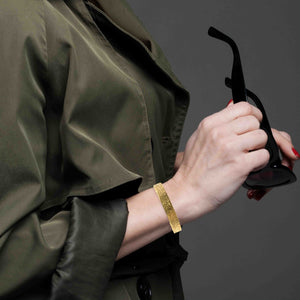 4 of 9: Model wearing a wide textured Nu Gold brass cuff bracelet on the wrist, holding sunglasses to show the bracelet’s scale, fit, and warm hammered gold finish.