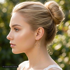 6 of 8: AI generated blonde model with hair styled in a bun wearing a 1 inch chased bronze hoop earring outdoors. The polished bronze hoop hangs from a French ear wire and is shown in side profile against a softly blurred green garden background and a light colored top.