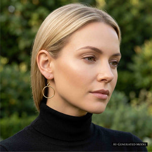 2 of 8: AI generated model with blonde hair tucked behind her ear wearing a 1 inch chased bronze hoop earring with a black turtleneck outdoors. The warm bronze hoop hangs from a French ear wire and stands out against the dark top and softly blurred green garden background.