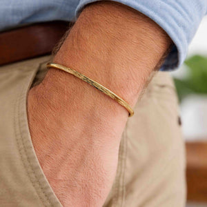 5 of 9: A close-up of a man's wrist wearing a 3.5mm bark-textured bronze cuff bracelet with a warm golden tone. The bracelet has a slim, open design and a natural, handcrafted finish. He is dressed casually in a light blue shirt and beige pants with a brown belt, showcasing the bracelet's understated and masculine style suitable for everyday wear.