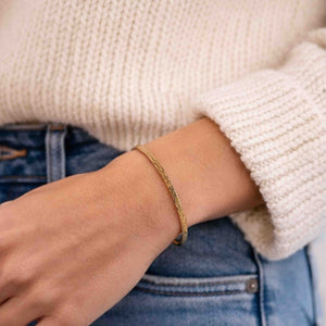 4 of 9: A close-up lifestyle photo of a woman wearing a slim 3.5mm bark-textured bronze cuff bracelet on her wrist. The bracelet shows a warm golden-bronze tone with a subtle organic texture. She is dressed casually in blue jeans and a soft cream knit sweater, highlighting the bracelet’s natural, everyday style and relaxed, modern look.