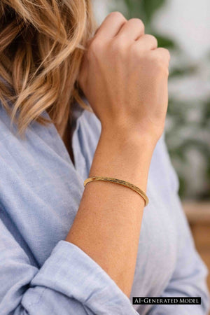 2 of 9: A woman wearing a handcrafted 3.5mm bark-textured bronze cuff bracelet on her wrist. The bracelet has a warm, golden tone and a slim, open design that rests comfortably on the skin. She is dressed in a light blue long-sleeve shirt, with her hand gently resting near her face, showcasing the bracelet’s natural, earthy elegance in a casual, everyday setting.  AI Generated Model.