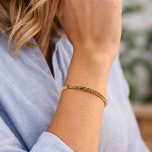 2 of 9: A woman wearing a handcrafted 3.5mm bark-textured bronze cuff bracelet on her wrist. The bracelet has a warm, golden tone and a slim, open design that rests comfortably on the skin. She is dressed in a light blue long-sleeve shirt, with her hand gently resting near her face, showcasing the bracelet’s natural, earthy elegance in a casual, everyday setting.