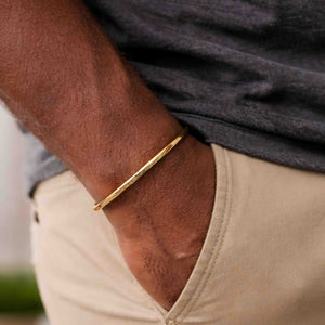 7 of 9: A close-up of a man's wrist wearing a slim 3.5mm bark-textured bronze cuff bracelet with a warm golden tone. The bracelet has a handcrafted, natural finish and a minimalist open design. He is dressed casually in a charcoal gray shirt and light beige pants, with one hand in his pocket, highlighting the bracelet’s relaxed and earthy everyday style.