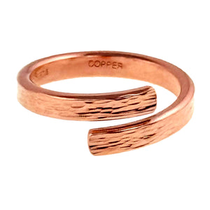 7 of 9: Angled side view of a slim solid copper bypass ring on a white background. The image shows the open wrap shape, smooth inner band, warm copper color, bark textured outer surface, and the copper stamp inside the ring.