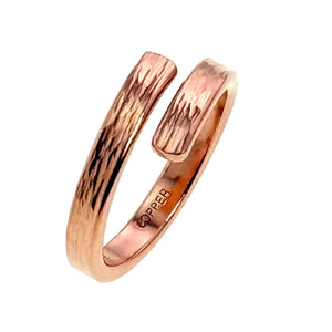 5 of 9: Vertical angled view of a narrow solid copper bypass ring on a white background. The ring curves upward to show both flat textured ends, the bark like surface marks, the open wrap design, and the copper stamp along the inner band.