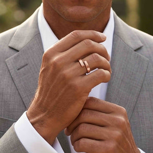 4 of 9: Man wearing a slim bark textured copper bypass ring while dressed in a light gray suit and white shirt. His hand is raised near his collar, showing the open wrap shape and two textured copper ends against a softly blurred outdoor background