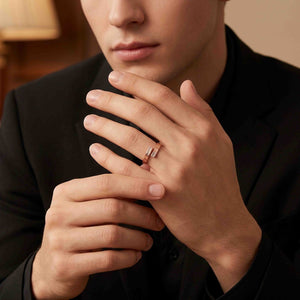 8 of 9: Man wearing a narrow copper bypass ring with bark texture while dressed in a black shirt and black jacket. His hands are held near his face, drawing attention to the warm copper tone and open wrap design in a softly lit indoor setting.