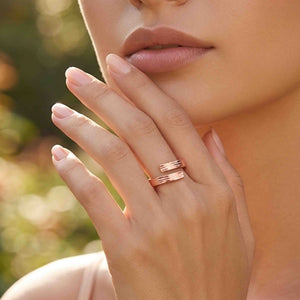 2 of 9: Close up of a woman wearing a slim bark textured copper bypass ring on her finger. The open wrap design ends in two flat textured tips. Her hand rests near her face against a softly blurred outdoor background with warm green and golden tones.