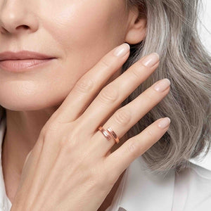 6 of 9: Close up of a woman with gray hair wearing a narrow copper bypass ring with a bark texture. The ring sits on her finger as she raises her hand near her cheek. A pale manicure and light clothing give the image a clean, polished look.