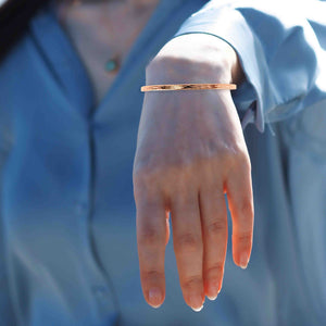 7 of 10: A close-up image of a woman's wrist wearing a slim, handcrafted 3.5mm wide copper cuff bracelet with a bark-textured finish. The bracelet has a warm reddish-gold hue and a smooth, polished surface that catches the light subtly. It rests above the wrist bone and stands out against the light blue satin blouse she is wearing. The background is softly blurred, creating a relaxed, outdoor atmosphere. The woman's hand is relaxed and facing downward, drawing attention to the bracelet's minimal yet natural design.