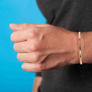 2 of 10: A close-up image of a person's wrist wearing a slim, handcrafted copper cuff bracelet with a bark-textured finish. The bracelet rests comfortably above the wrist bone, showcasing its thin 3.5mm width and natural reddish tone. The copper’s raw, polished surface highlights the subtle organic bark pattern. The person is wearing a dark shirt, and the background is a bright blue, creating a clean contrast that draws attention to the bracelet’s warm metallic sheen and minimalist design. The bracelet’s open-ended 