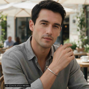 6 of 10: AI generated male model seated at an outdoor cafe wearing a gray shirt and slim brushed stainless steel cuff bracelet. The bracelet is visible on his wrist as he rests his hand near his face, showing how the narrow silver tone design looks with casual clothing.