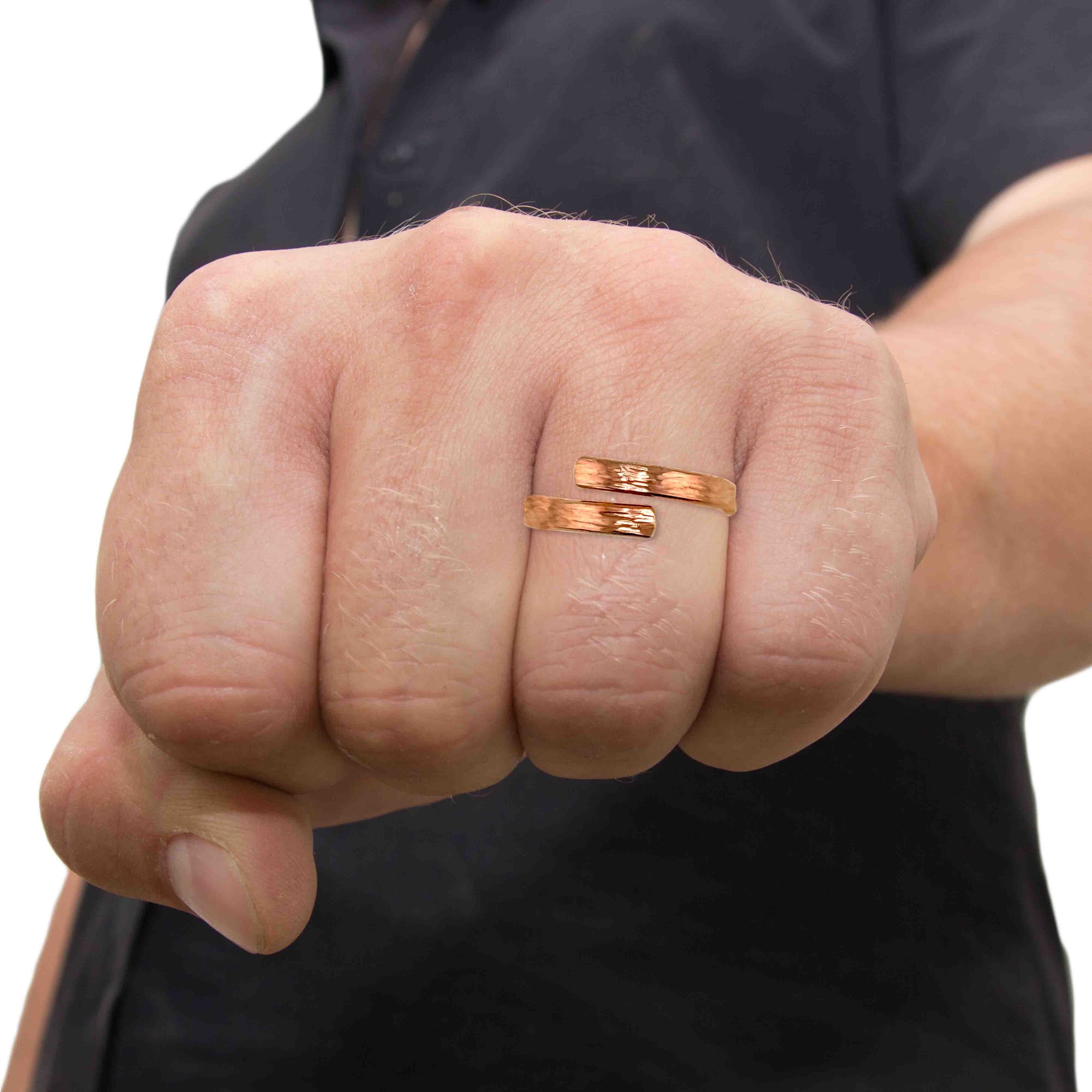 Close-up of a hand wearing a solid 3.5mm Chased Copper Bypass Ring with an open wrap design. The ring curves around the finger with two flat ends that nearly meet at the front, creating a clean modern silhouette. Its slim band has a lightly chased texture that reflects the light, adding subtle detail to the warm copper finish. The ring is shown on a clenched hand against a dark shirt background, emphasizing its bold yet minimalist look.