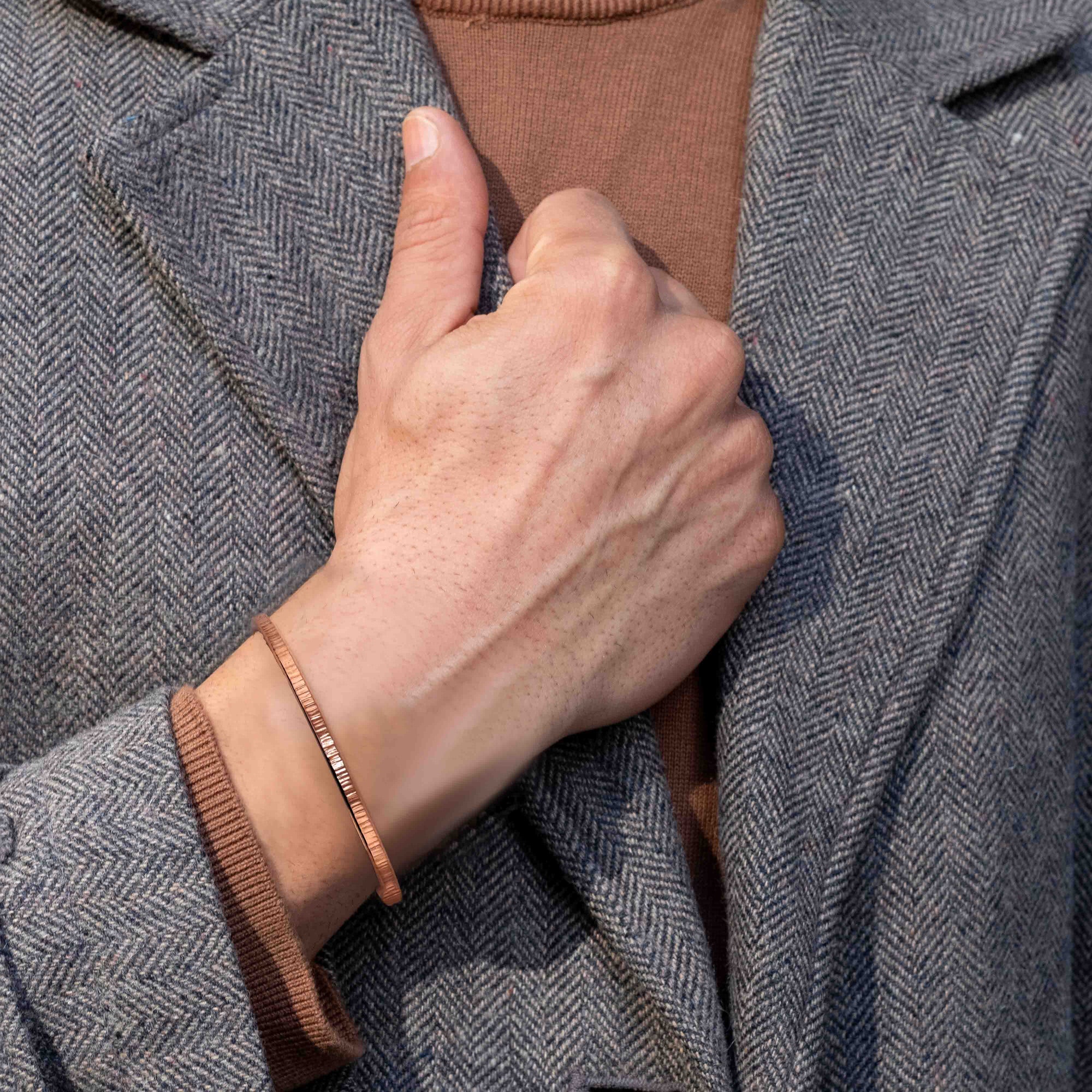 Close-up of a person’s hand gripping the lapel of a gray herringbone coat, showing a slim chased copper cuff bracelet worn over the cuff of a brown knit sweater.