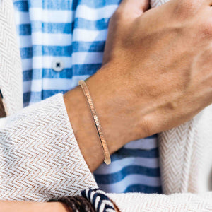 8 of 11: A man wearing a slim, hand-textured copper cuff bracelet paired with a blue and white striped shirt and a cream herringbone jacket, highlighting a stylish and casual look.