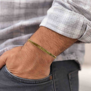 6 of 9: Close-up of a man's wrist wearing a 3.5mm chased Nu Gold brass cuff bracelet with a lightly textured, ridged finish. The bracelet has a warm golden tone and minimalist design. He is dressed in a light gray plaid shirt with dark jeans, and his hand is casually tucked into his pocket, showcasing the bracelet's laid-back yet refined look.