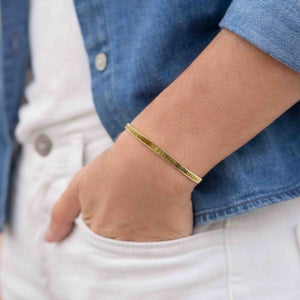 8 of 9: Close-up of a woman's wrist wearing a 3.5mm chased Nu Gold brass cuff bracelet with a subtle ridged texture and warm golden tone. The hand is casually placed in the pocket of white pants, paired with a light denim jacket, creating a relaxed, stylish look. The bracelet’s minimal design adds a touch of shine, making it an easy everyday accessory.
