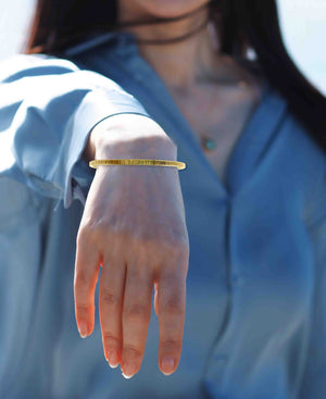 2 of 9: Woman wearing a slim 3.5mm chased Nu Gold brass cuff bracelet on her wrist, shown in natural daylight. The bracelet has a warm golden tone and a subtle textured finish that reflects light. She is dressed in a light blue blouse, and her hand is relaxed and extended downward, highlighting the bracelet’s delicate yet stylish appearance. The photo captures a casual, elegant look suitable for everyday wear.