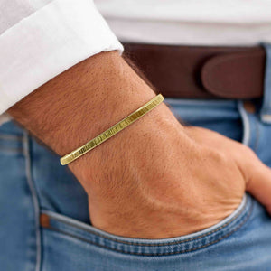 4 of 9: Close-up of a man's wrist wearing a slim 3.5mm chased Nu Gold brass cuff bracelet with a subtle ridged texture. The bracelet has a warm golden tone and minimalist design, complementing the casual outfit of a white shirt, brown belt, and blue jeans. The man's hand is relaxed and resting in his pocket, showcasing the bracelet's understated, everyday style.