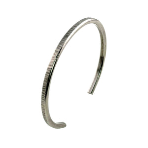 8 of 11: Angled left side product view of the John S. Brana 3.5mm Chased Stainless Steel Cuff Bracelet standing upright against a white background. The photograph highlights the curved open cuff shape, narrow 3.5mm width, and 2.5mm thick construction. Hand chased marks follow the outer surface of the silver tone 316L surgical stainless steel, contrasting with the smooth interior and rounded bracelet ends.