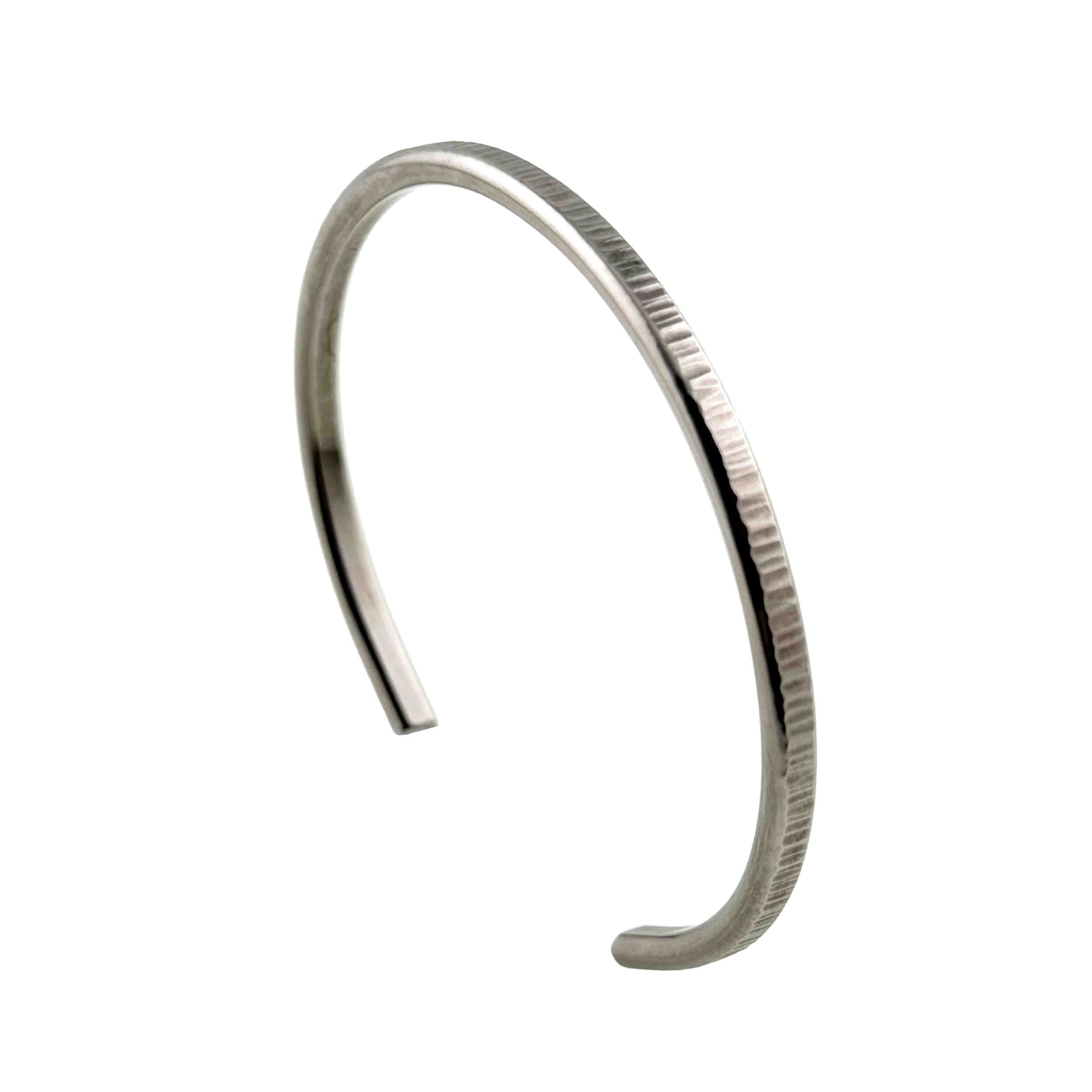 Main product photograph of the John S. Brana 3.5mm Chased Stainless Steel Cuff Bracelet displayed upright at an angle on a white background. The handcrafted bracelet features a slim, curved open cuff design made from hypoallergenic 316L surgical stainless steel. Fine vertical chased marks decorate the outer surface, creating a textured silver tone finish, while the smooth interior and rounded ends complete the minimalist design.