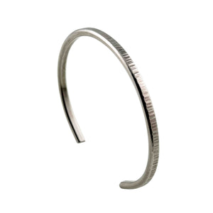 1 of 11: Main product photograph of the John S. Brana 3.5mm Chased Stainless Steel Cuff Bracelet displayed upright at an angle on a white background. The handcrafted bracelet features a slim, curved open cuff design made from hypoallergenic 316L surgical stainless steel. Fine vertical chased marks decorate the outer surface, creating a textured silver tone finish, while the smooth interior and rounded ends complete the minimalist design.