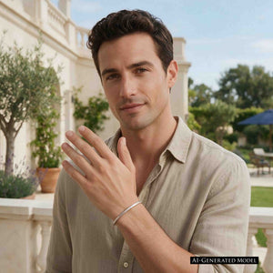 9 of 11: AI generated male model with short dark hair wearing a beige collared shirt and the John S. Brana 3.5mm Chased Stainless Steel Cuff Bracelet. He raises his hand near his face, showing the slim silver tone bracelet on his wrist. The handcrafted 316L surgical stainless steel cuff features a textured chased finish and minimalist design, photographed in a sunny outdoor courtyard with greenery.
