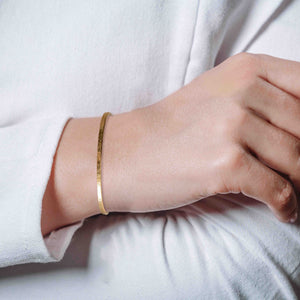 8 of 10: Close-up lifestyle image of a slim hammered bronze cuff bracelet worn on a woman’s wrist. The narrow 3.5mm solid bronze cuff features a warm golden tone and a subtle hand-hammered texture that reflects light gently. The open cuff design curves smoothly around the wrist for a minimalist look. The bracelet is styled with a soft white long sleeve top, creating a clean, simple, and modern feel.