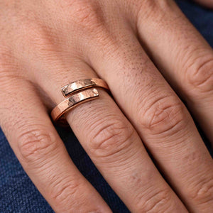 2 of 7: A close-up view of a hand wearing a 3.5mm hammered copper bypass ring on the middle finger. The ring features an open front design with two polished, textured ends that curve past each other. The warm reddish tone of the raw copper contrasts subtly against the natural skin tone. The hammered finish catches the light, giving the ring a rugged yet refined look. Blue denim fabric is partially visible in the background.