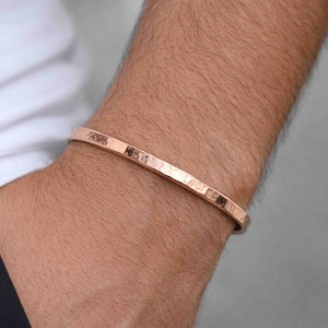 2 of 9: Close-up of a person’s wrist wearing a slim 3.5mm hand-hammered copper cuff bracelet. The raw copper surface shows a warm reddish shine with subtle hammered texture catching the light. The open-ended cuff fits snugly around the wrist for a minimalist look. A light sleeve edge appears near the wrist, and the background is softly blurred in neutral gray tones.