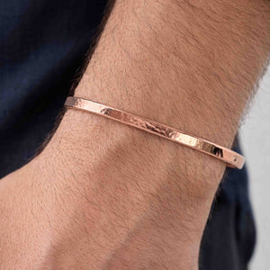 6 of 9: Close-up of a man wearing a 3.5mm hand-hammered copper cuff bracelet on his wrist. The bracelet has a warm, polished copper finish with a textured surface that reflects light. Its slim, open-ended design curves gently around the wrist for a comfortable fit. The person is wearing a dark blue shirt, and the bracelet stands out against their skin tone, highlighting its handcrafted, minimalist style.