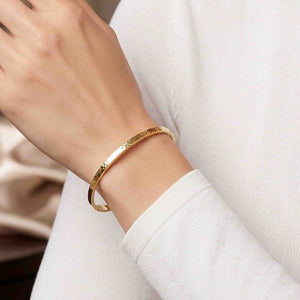 4 of 10: Close-up of a woman wearing a slim hammered Nu Gold brass cuff bracelet on her wrist, styled with a soft white long sleeve top. The narrow 3.5mm cuff has a lightly textured surface that reflects warm golden tones. Her hand is gently raised, showing a clean, minimal look in a relaxed indoor setting with soft, neutral background details.
