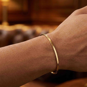 7 of 10: Close-up of a slim hammered Nu Gold brass cuff bracelet worn on a wrist, highlighting its narrow 3.5mm width and softly textured surface. The open cuff sits just above the wrist bone, reflecting warm golden tones against the skin. The background is softly blurred with warm brown hues, giving a cozy, lifestyle feel that emphasizes the bracelet’s simple, handcrafted style.