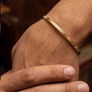 2 of 10: Close-up of a slim hammered Nu Gold brass cuff bracelet worn on a wrist, showing its narrow 3.5mm width and lightly textured surface that reflects warm golden tones. The open cuff design sits comfortably above the wrist bone, styled on skin with natural texture visible, highlighting its minimalist, handcrafted look in a lifestyle setting.